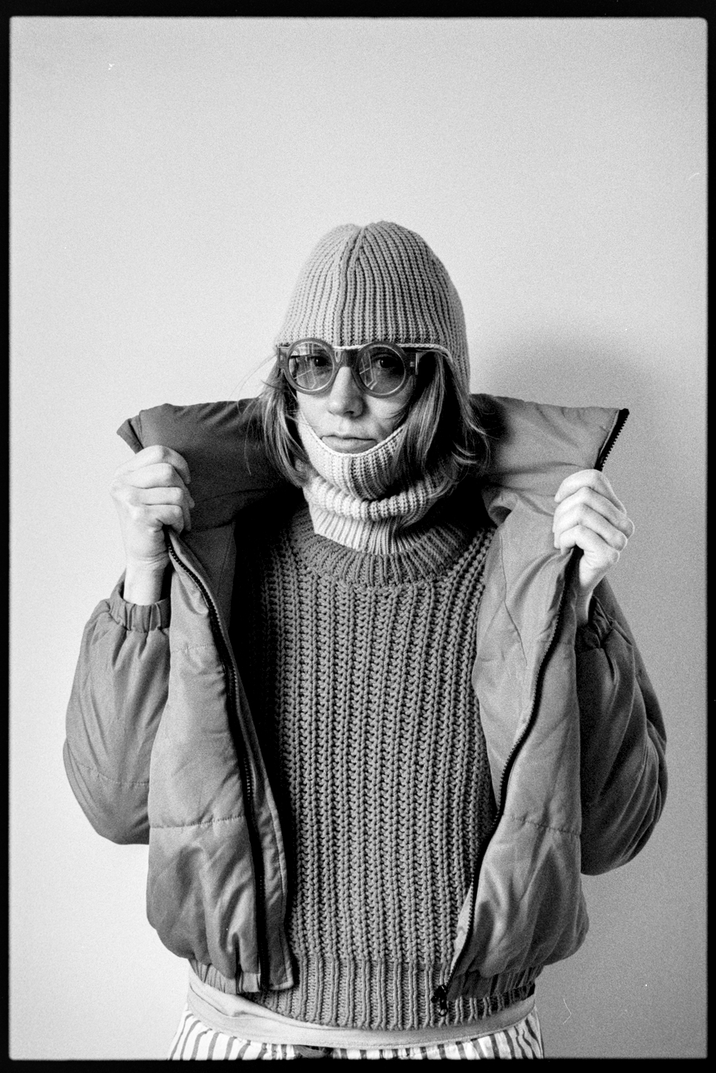 Kallie in a ski mask and round sunglasses holding open her snow jacket