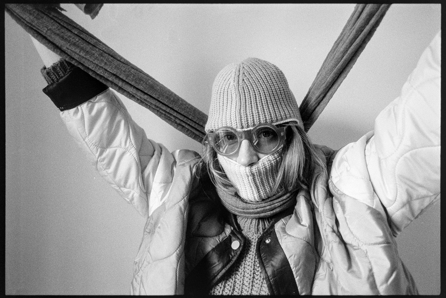 Kallie in a ski mask and round sunglasses holding her scarf up over her head