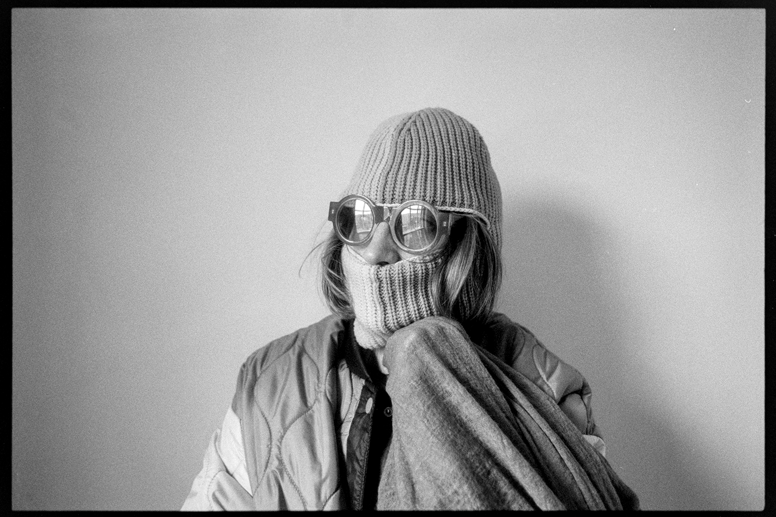 Kallie in a ski mask and round sunglasses holding her scarf to her chin and staring out the window with the glare in her glasses