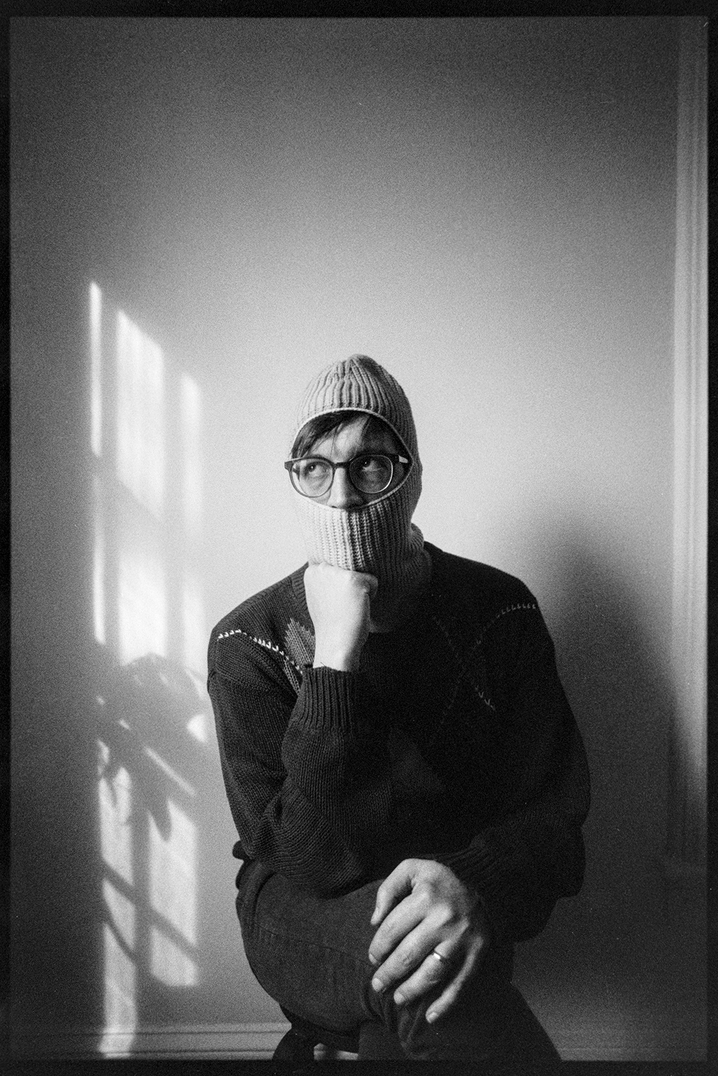 Jason in a ski mask with his hand on his chin, the other hand on his knee, looking to the side toward the window