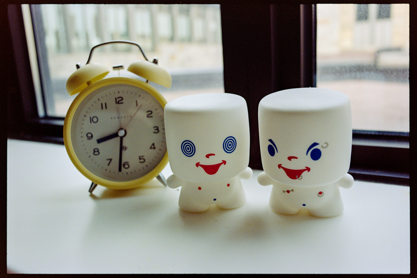 A yellow alarm clock next to two plastic marshmallow men toys