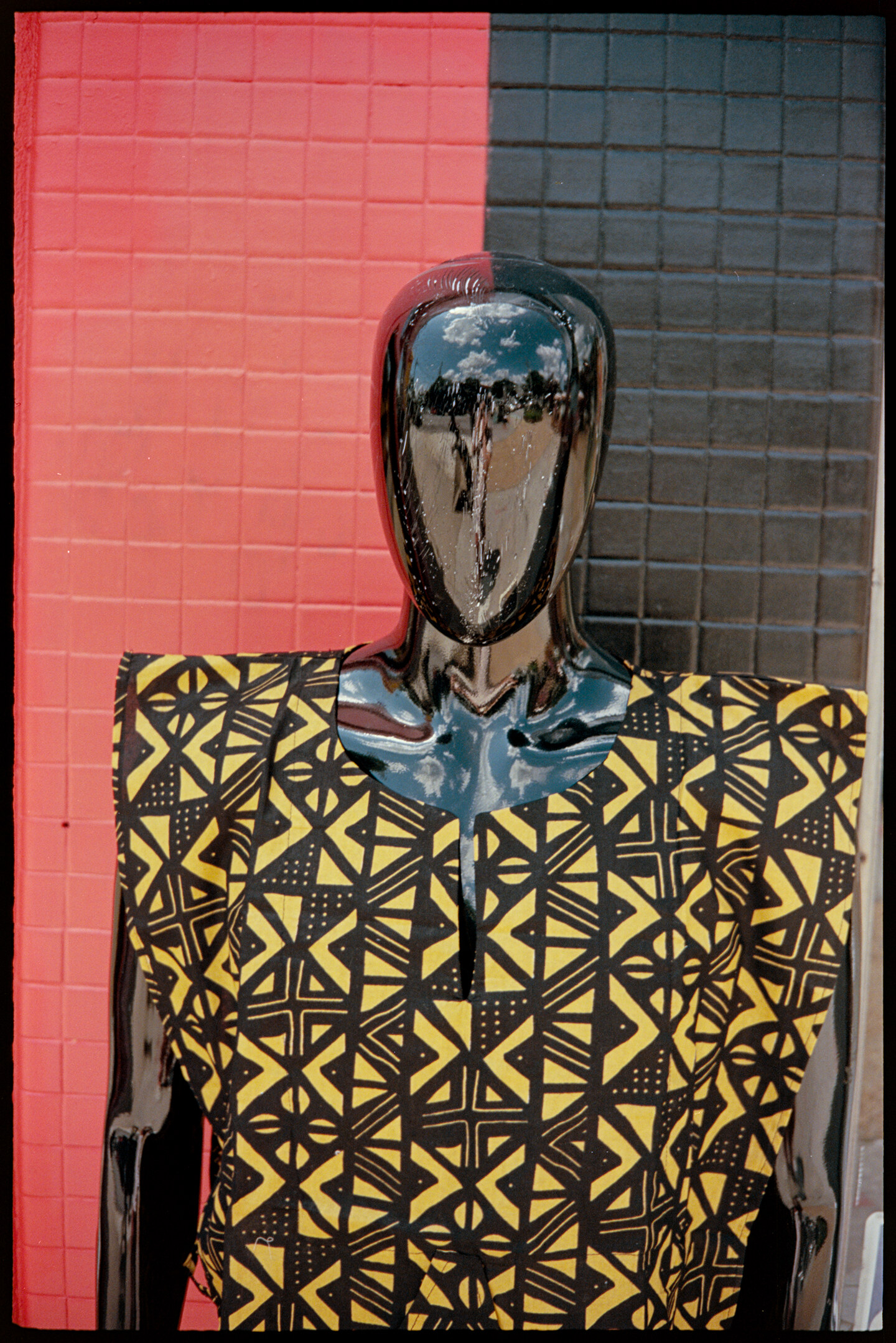 A black mannequin wearing a yellow African shirt in front of a shop building with red and black tile on the wall in Atlanta