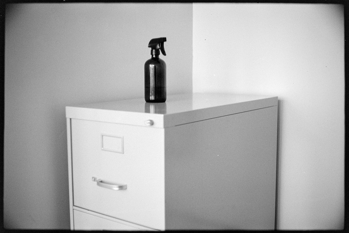 A dark glass spray bottle sitting on top of a filing cabinet in the corner of an office