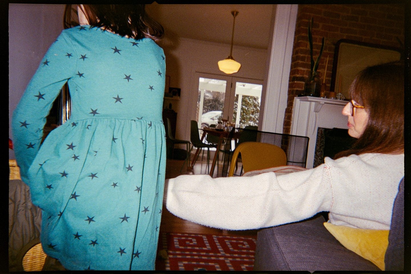 A woman sitting on a couch reaching her arm out to her young daughter in a teal dress with dark stars on it, shot from behind them.