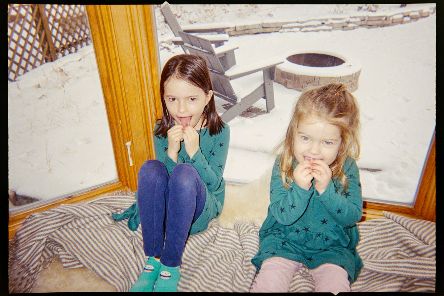 Two young sisters sitting in a bay window in a kitchen eating snacks and looking into the camera.