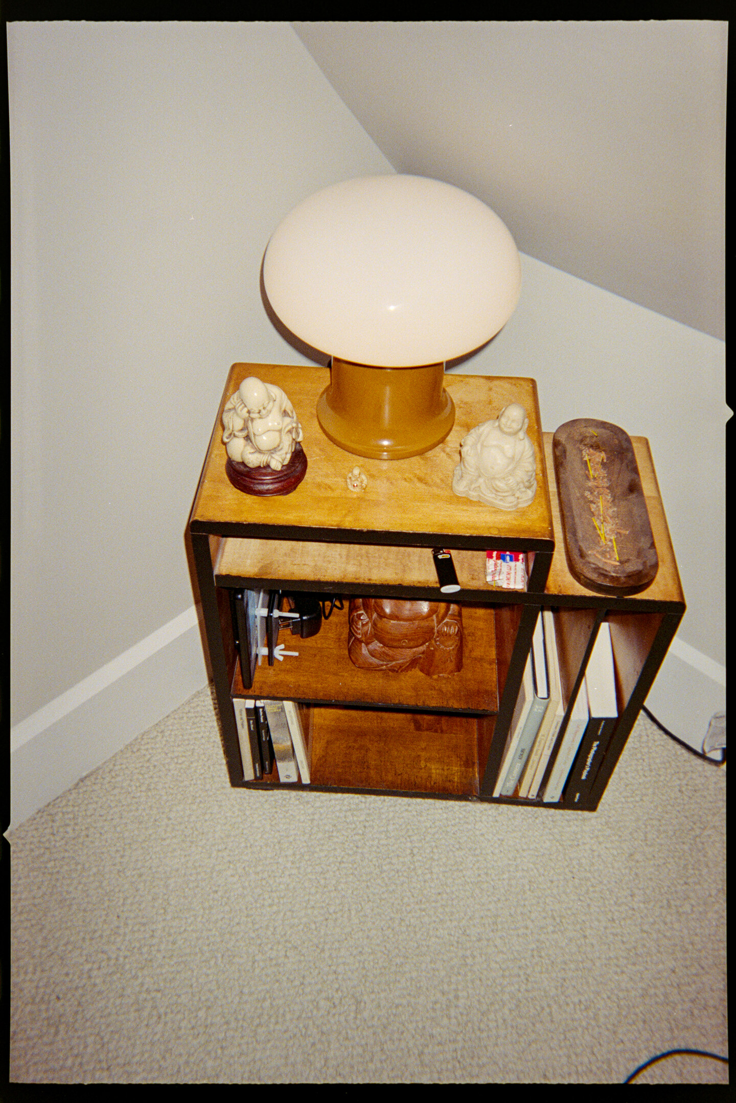 A night stand in a corner with a lamp, two small Buddhas, and books on it.