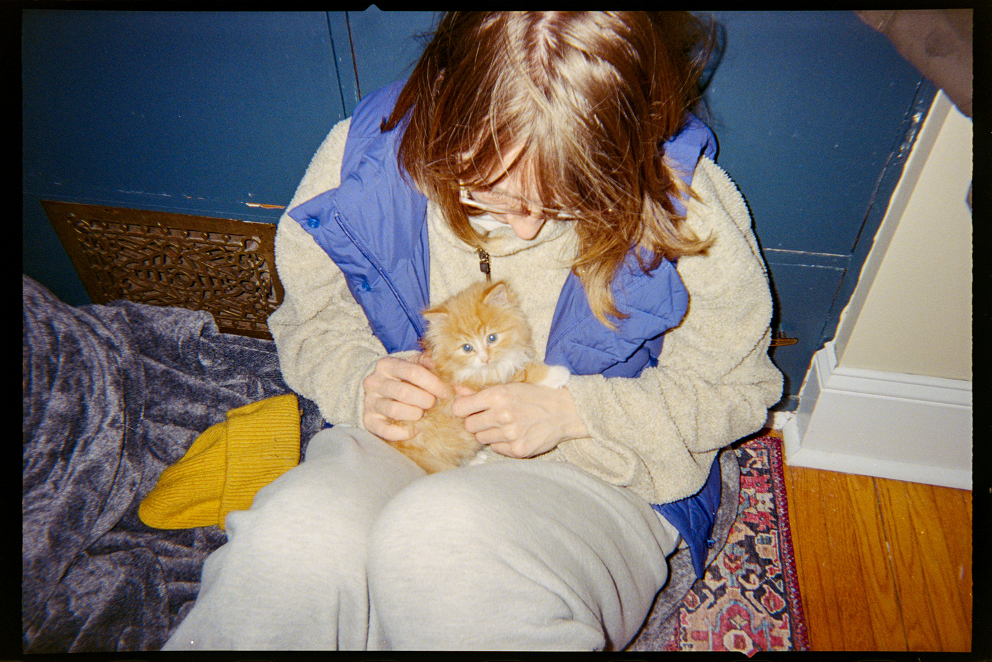 A woman in a blue vest holding a tiny orange kitten in her lap.
