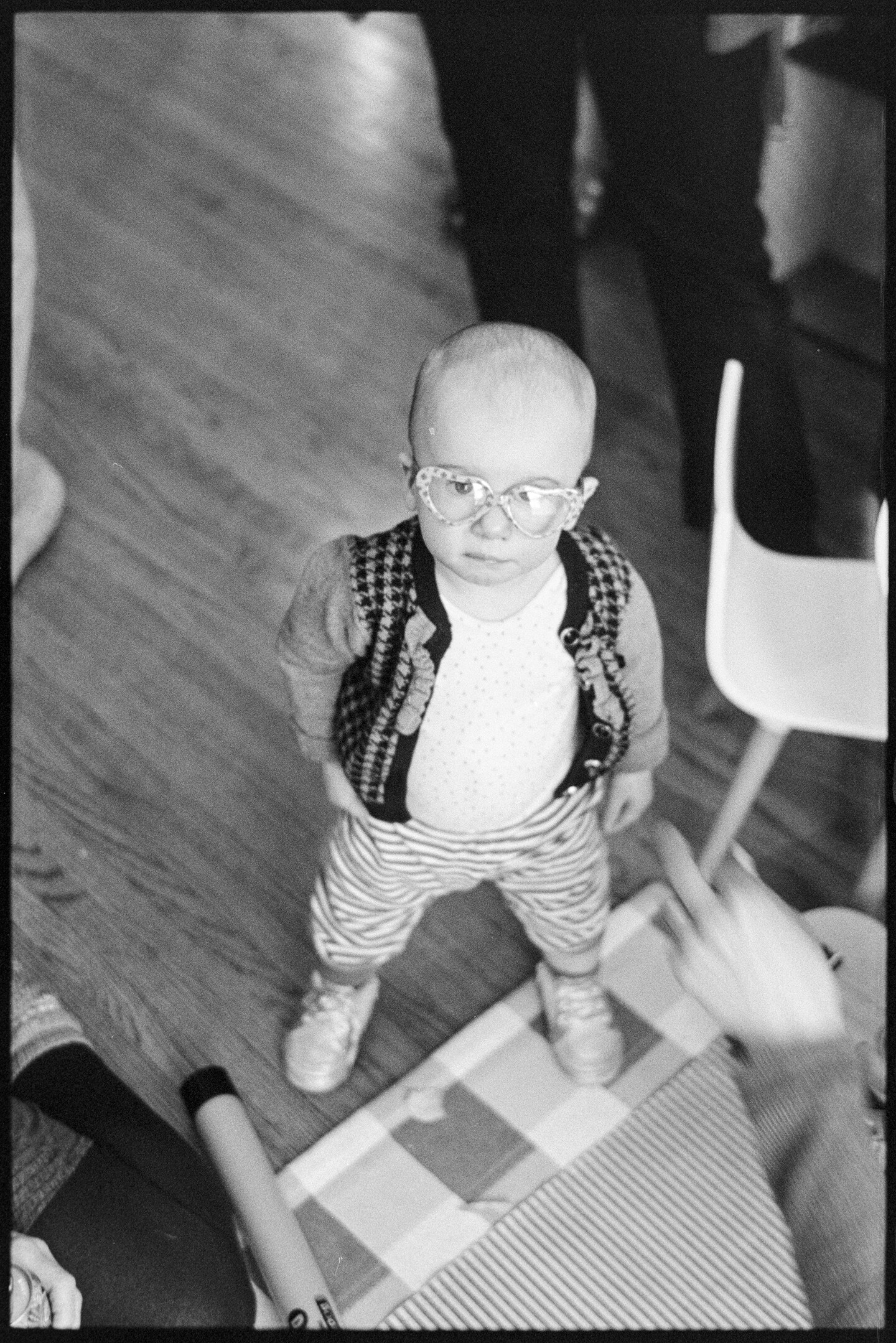 A toddler girl standing on the floor at a party surrounded by adults, looking up into the camera