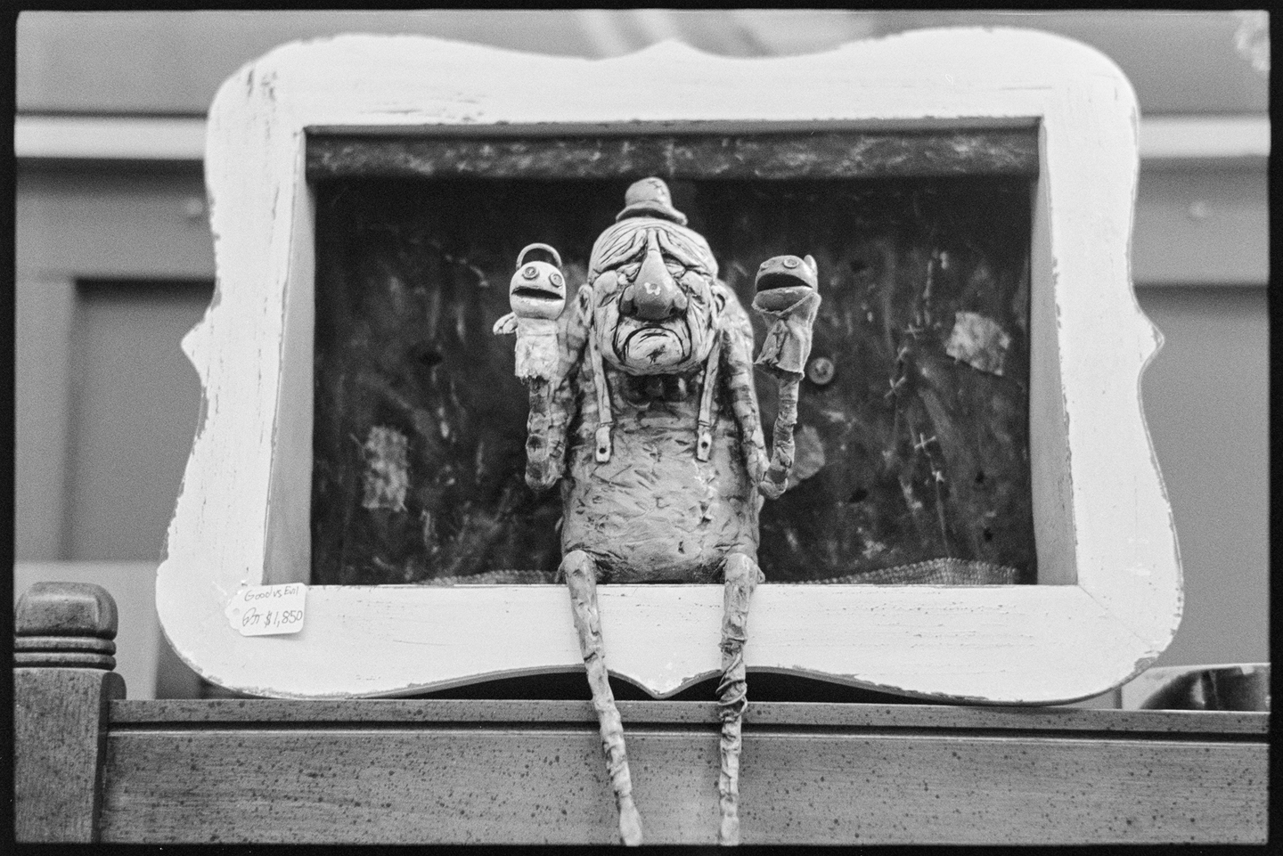A small clay puppeteer with a frowning face holding two puppets on his hands, set in a frame sitting on the shelf of an antique store