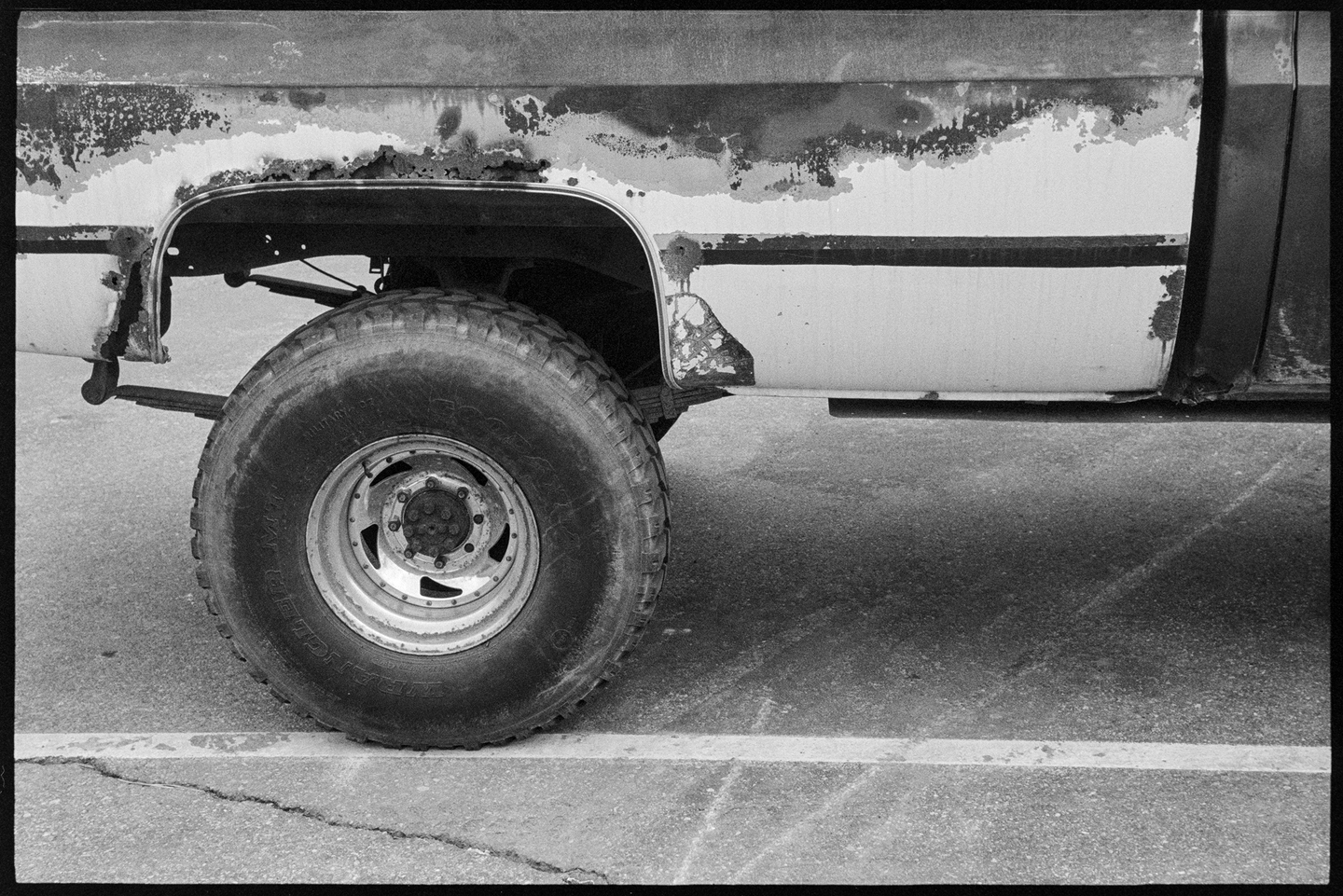 A side view of an old rusted pickup truck parked on the street, only the back tire and truck bed are in frame