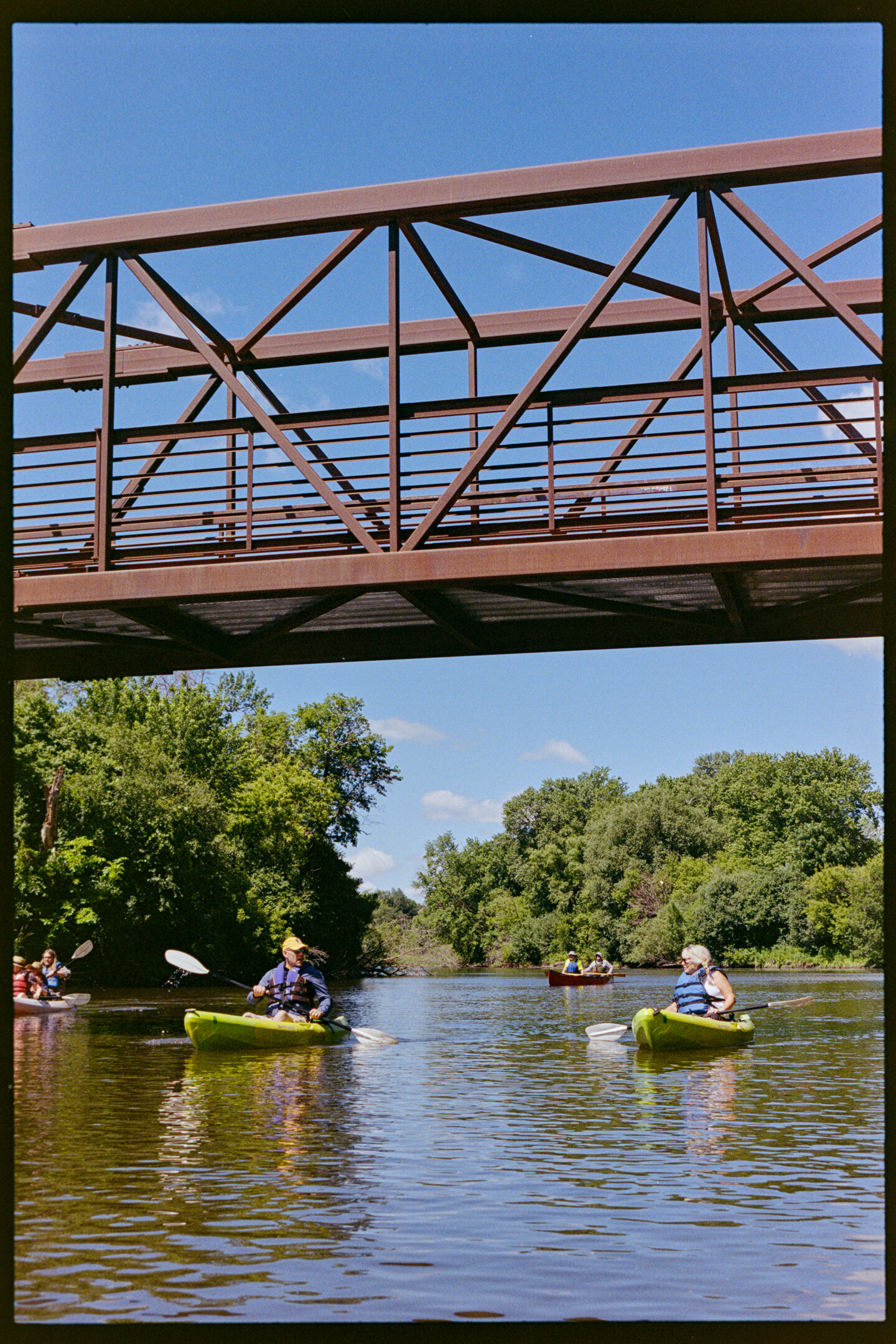 Kayakers passing under a rust-colored bridge on the Cannon River