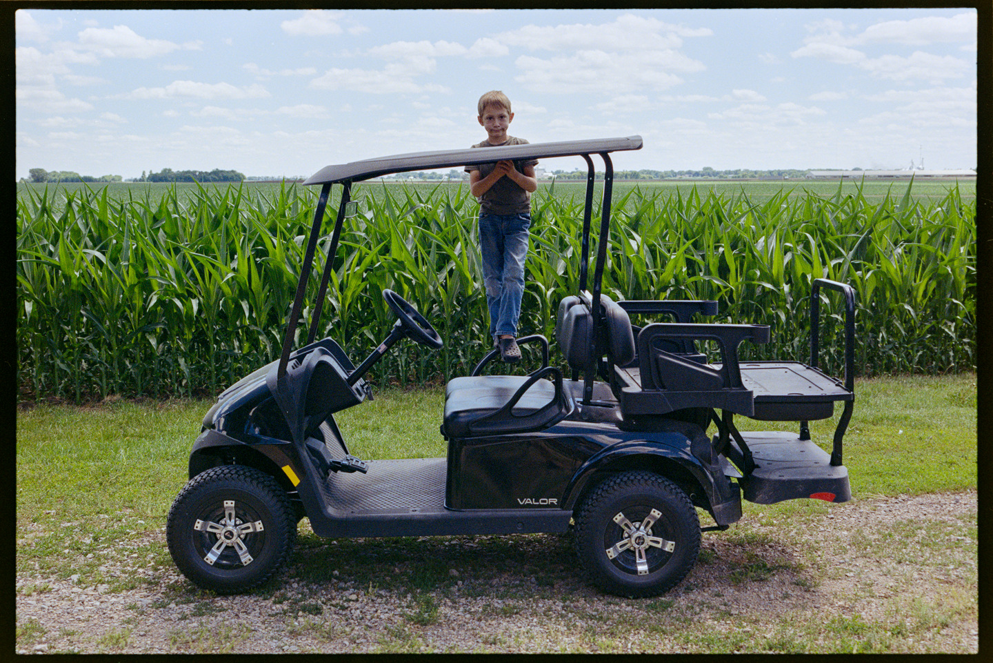 A young boy standing on a golf cart on the farm with a bright green corn field behind him