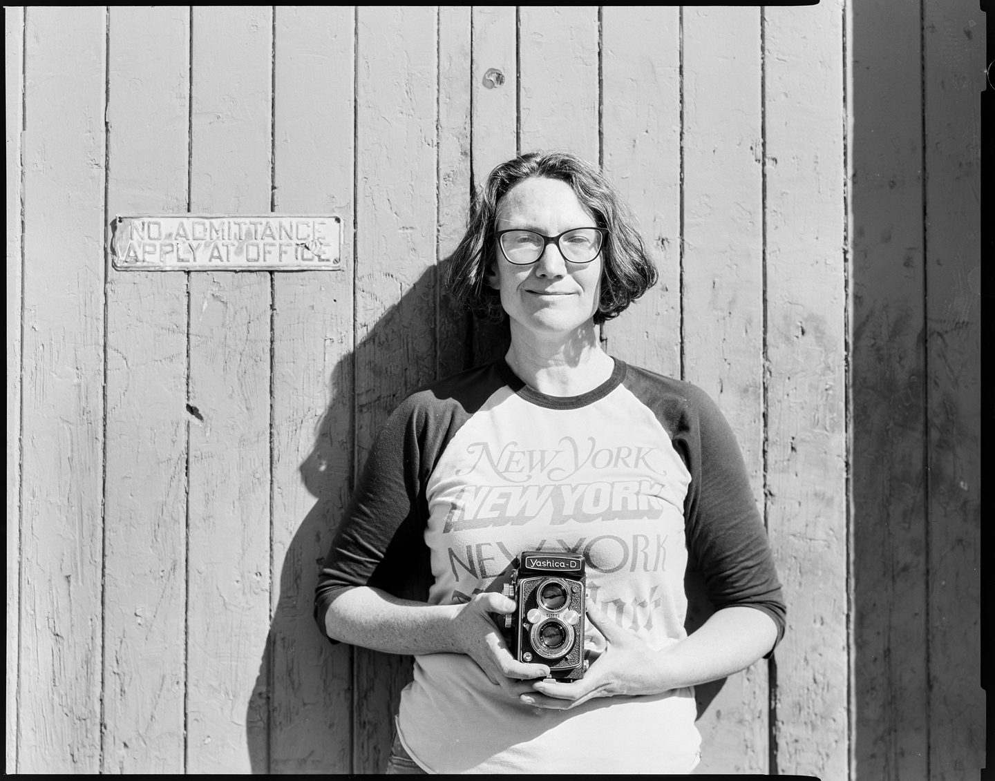 A woman standing in front of an old garage door holding a Yashica TLR camera. A sign on the door says No Admittance Apply at Office.